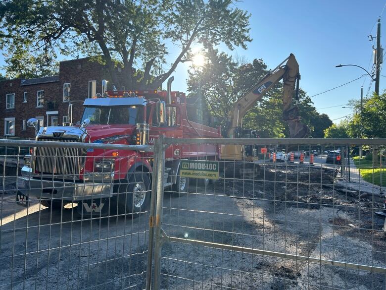 A construction site blocks access to two roads. A large red dump truck is parked on an unpaved stretch of the road. An excavator is parked behind it. The morning sun is peering out from some trees.  