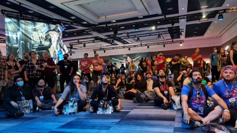 Dozens of people are seen standing and sitting on the floor, looking at a projection screen off-camera, in a large convention hall.