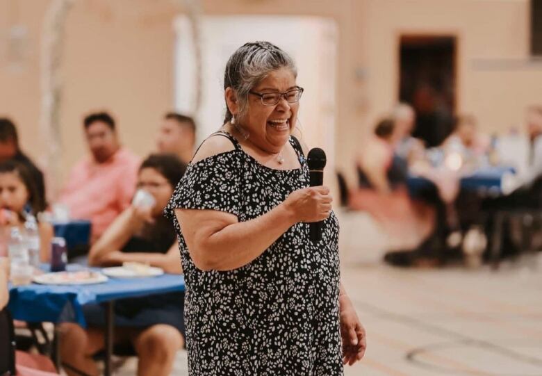 A smiling woman with a black and grey braid holds a microphone at a wedding in a gymnasium.