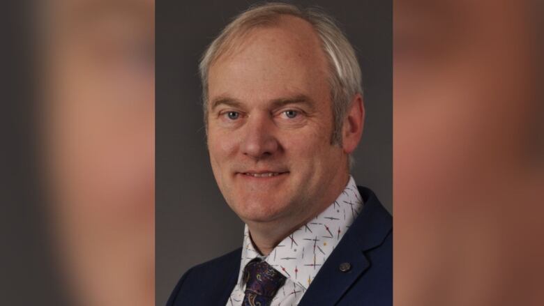 A man's headshot is pictured. He is wearing a tie and has grey hair.