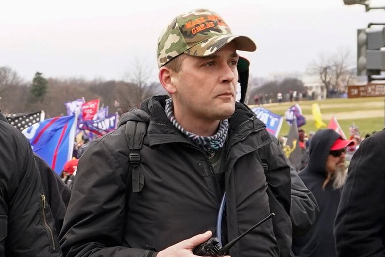 Man in camo cap and dark jacket, holding what looks like a walkie talkie, infront of a gathered group of people with flags and banners. 