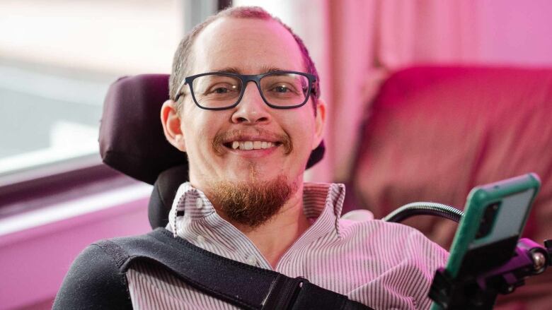 A person with a beard and glasses sits in a power wheelchair which has an attachment arm holding a phone in front of them. They are smiling.