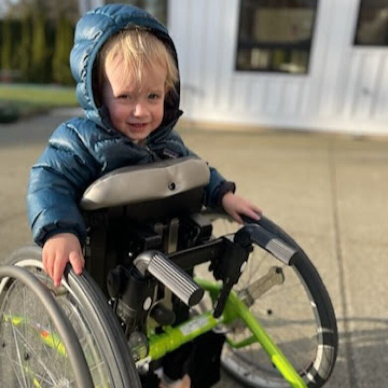 A child wearing a puffer jacket is smiling and standing with a walker with large wheels on either side.