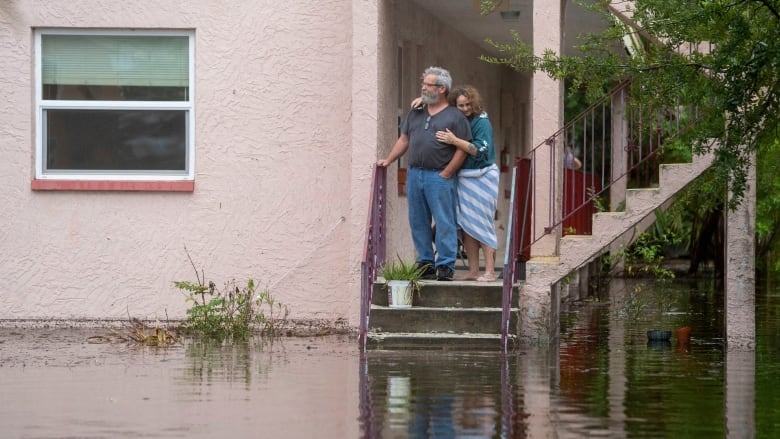 A woman embraces a man from behind as they stand looking out from a porch, surrounded by flood water