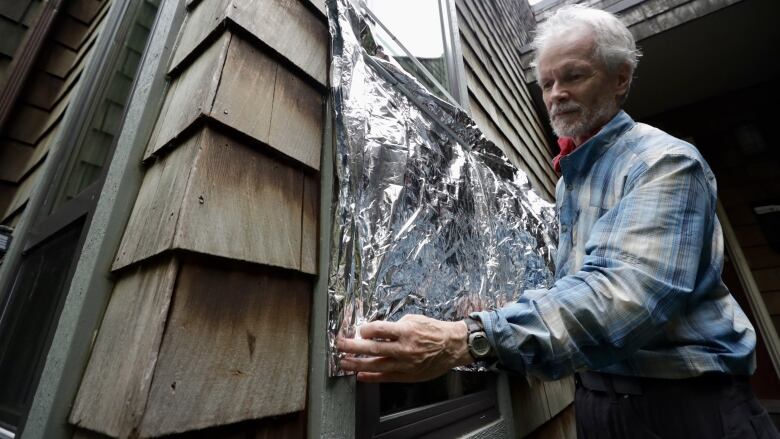 A man in a blue plaid shirt uses tin foil to cover a window. 