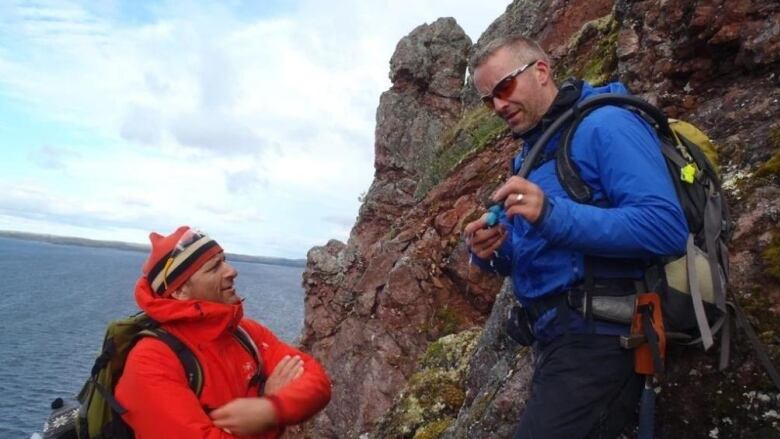 Two men are seen looking at a rock near the edge of a cliff overlooking water