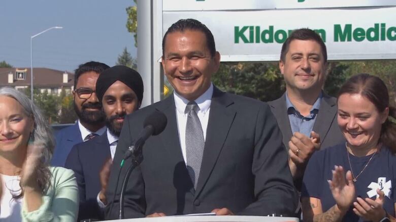 A politician in a grey suit makes a campaign announcement flanked by colleagues outside a hospital.