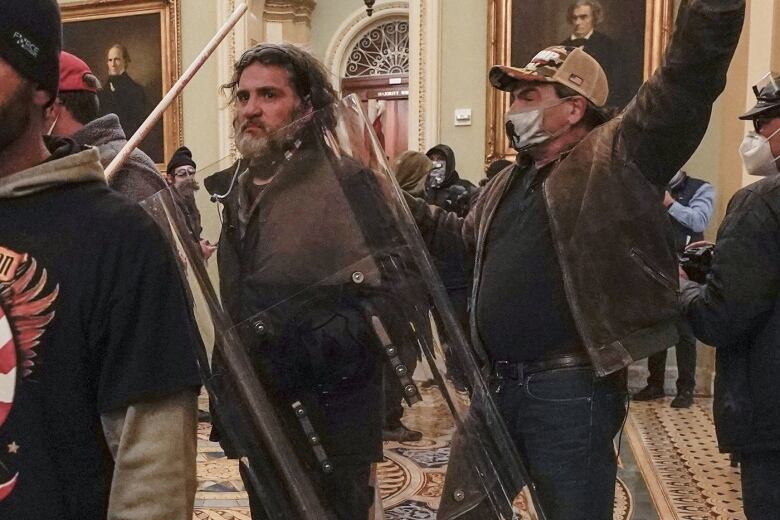 One man carries a police riot shield while another man raises his fist in the air while walking through a large, ornate room. 