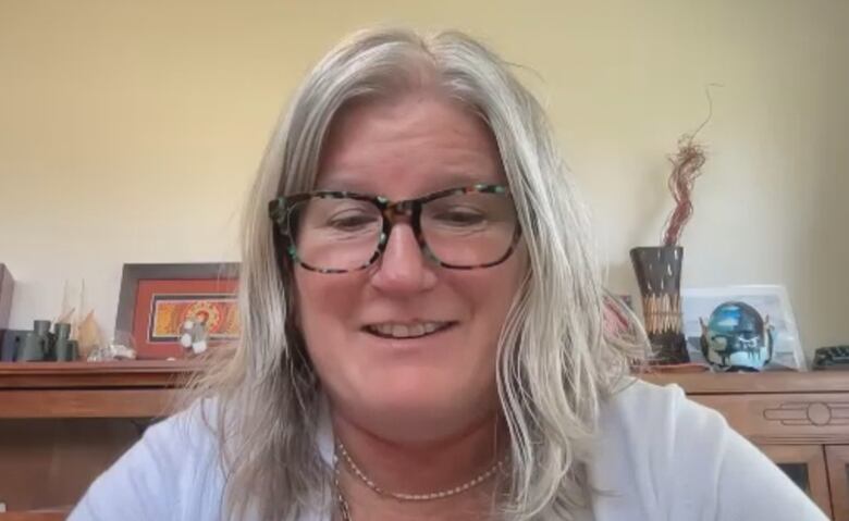 A woman with grey hair faces the camera in a Zoom interview, a bookcase and ornaments on the wall behind her.