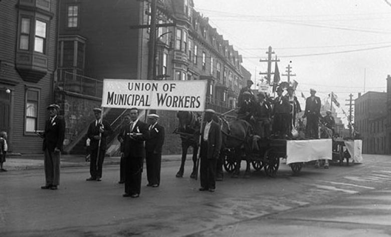 An old black and white picture of a parade of men and floats. Two men carry a banner that reads 