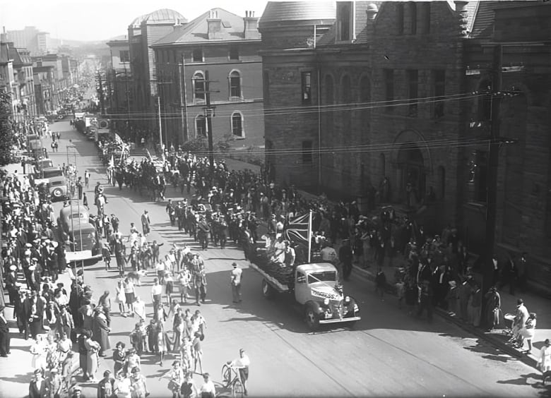 An overhead view in a black and white picture of a parade of men and cars on a 1930s city street. Spectators line both sides of the street.