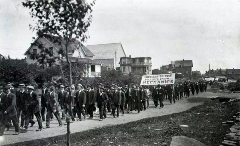 A black and white image of a line of men in dark suits marching down a town road.