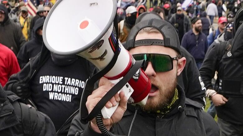 A man with sunglasses speaks into a megaphone. There is a large crowd behind him.