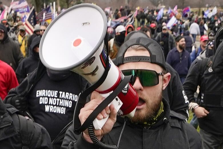 A man with sunglasses speaks into a megaphone. There is a large crowd behind him.