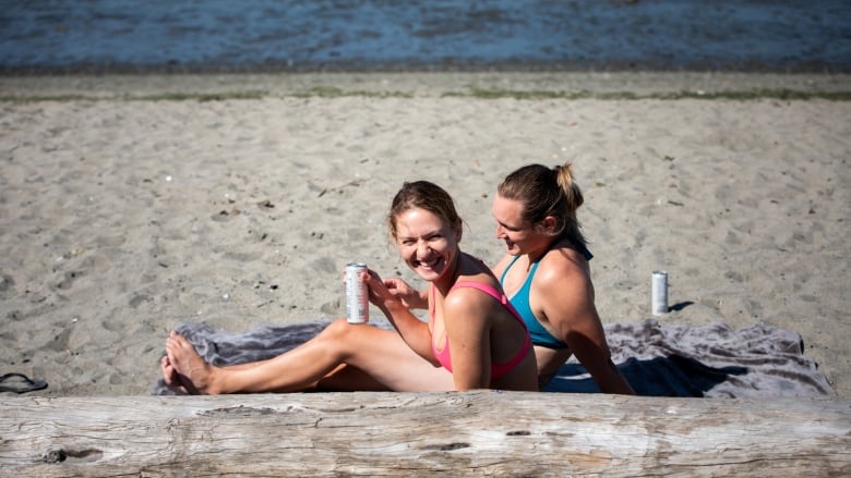 Two women in bikinis smile and drink alcoholic beverages at Spanish Banks beach.