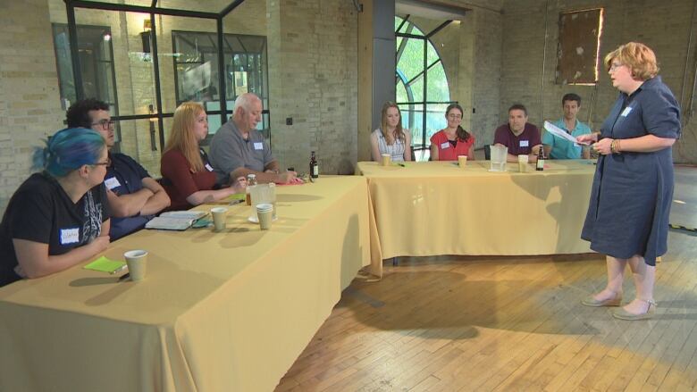 Eight people sit at two connected tables, while listening to a woman who is standing.