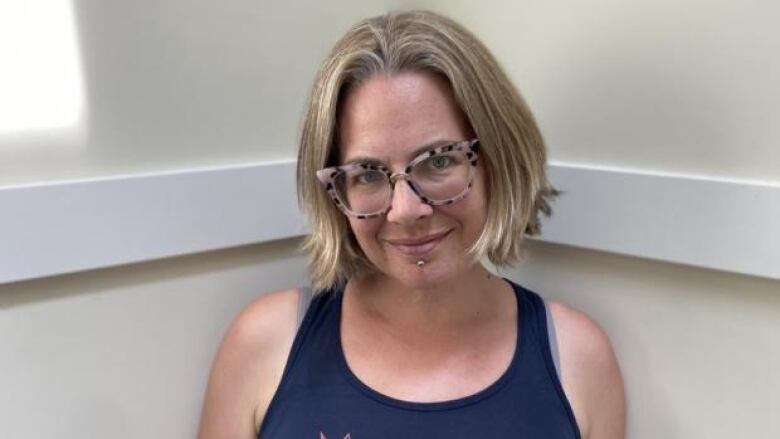 A woman with short sandy blonde hair and pink glasses with a piercing under her mouth and wearing a blue tanktop, she is smiling and looking straight at the camera