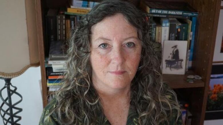 A woman in front of a wooden bookshelf next to a lamp. She has long curly grey hair and freckles on her face.