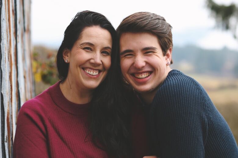 A woman in a pink sweater smiles besides a teenage boy, also smiling. 