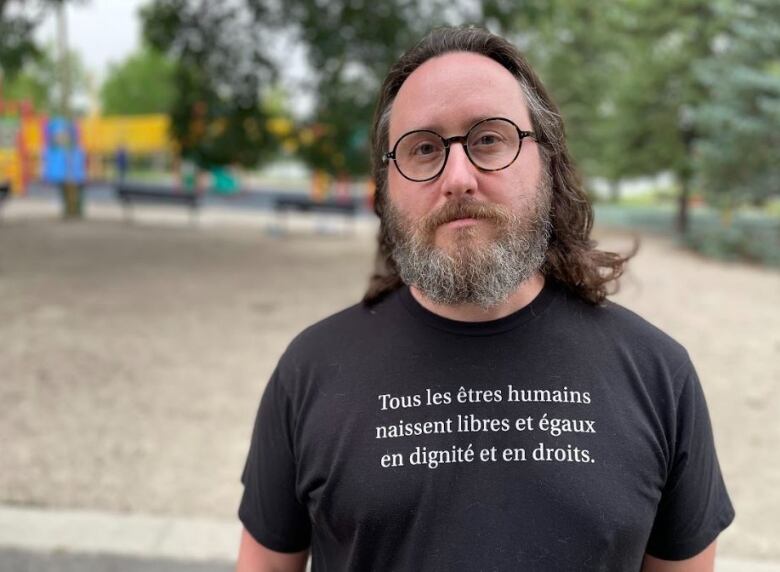 Man with shoulder-length graying brown hair and glasses poses for photo in front of playground in a black T-shirt.