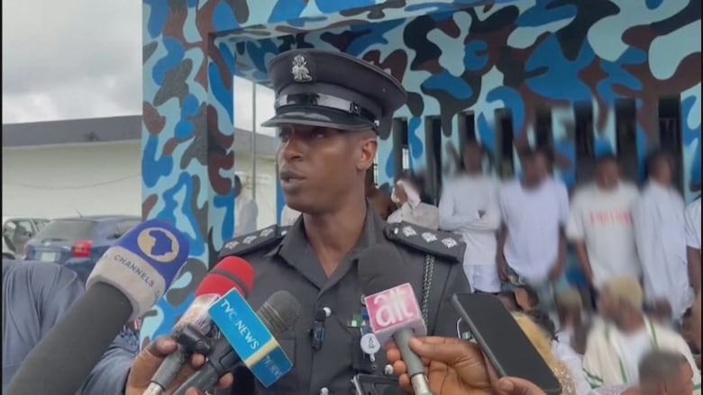 A man in a police uniform stands in front of microphones. Behind him are rows of people whose faces have been blurred.