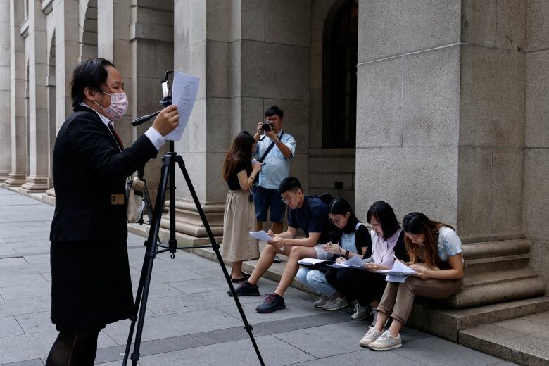 People sit and stand in front of a building as they read over some papers.