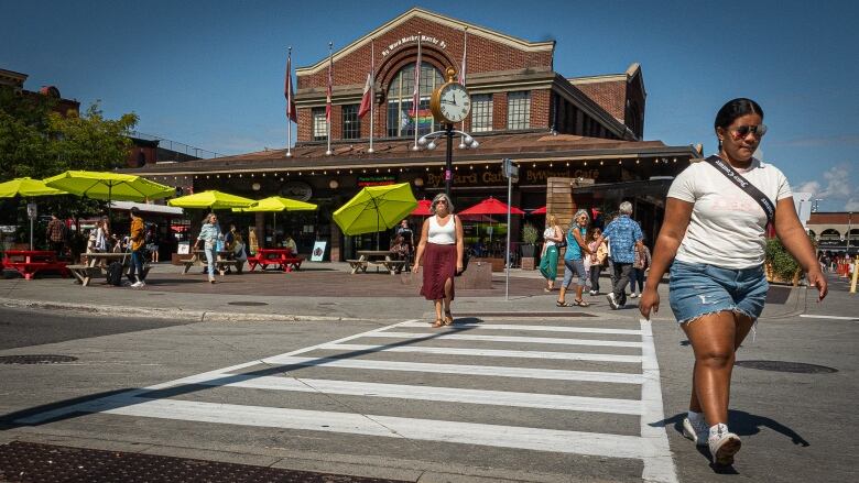 People walk around the outside of a heritage city market building on a summer day.