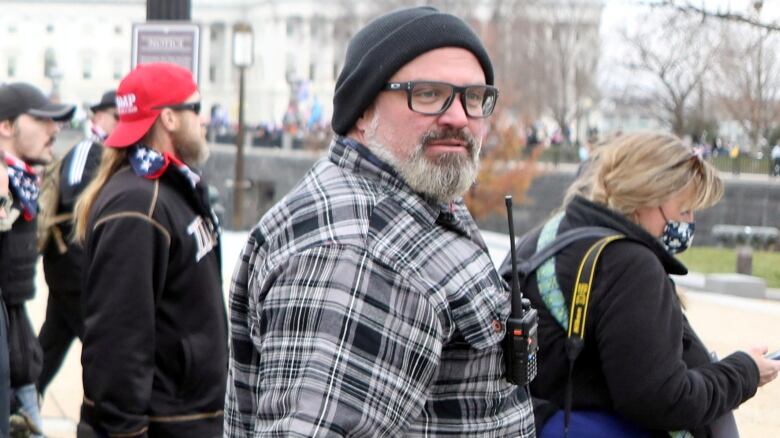 Man with grey beard and mustache, wearing a grey and white plaid coat and black tuque, walks by  