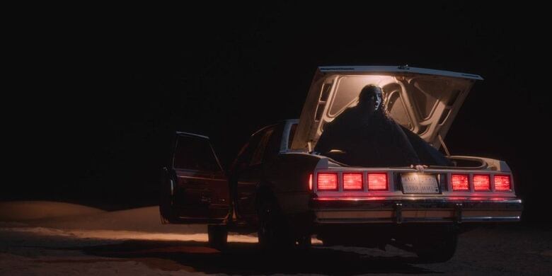 Still from NAGA. Nighttime image. A car with its lights on illuminates the pitch black sky. A young woman sits up in the car's open trunk, an expression of shock on her face.