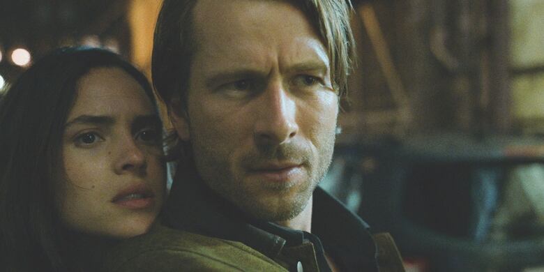 Still from Hit Man. Closeup of two actors, Adria Arjona (left) and Glen Powell. Adria rests her chin on Glen's shoulder. They appear to be peering cautiously at something off screen.