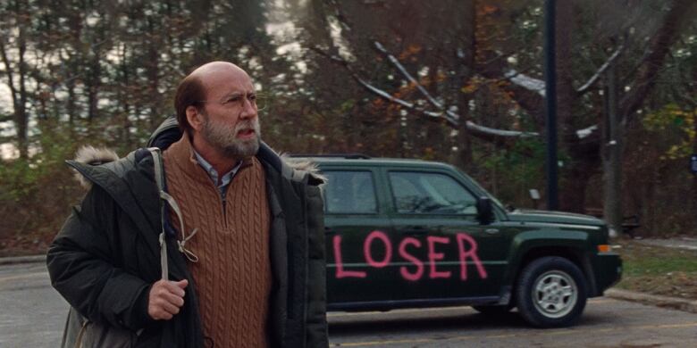 Still from Dream Scenario. Cloudy day. Nicolas Cage appears at left, bald and bearded, wearing a sweater and open parka. He is in a parking lot. A car appears behind him. Its side door has been spraypainted in pink. Text reads: 