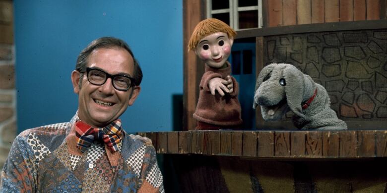 Archival photo of Mr. Dressup, Casey and Finnegan, gathered around the show's treehouse set. Mr. Dressup, a smiling man in dark glasses and a patchwork shirt, appears to be dressed in a '70s style. At his right is a puppet stage in the shape of a child's treehouse. Hand puppets Casey -- a freckled red headed child with black spots for eyes wearing a turtleneck -- and Finnegan -- a fuzzy grey dog with floppy ears -- rise from the treehouse porch.