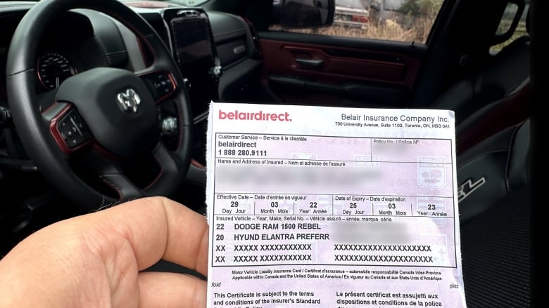 A person holds up a car insurance document with identifying information blurred out in front of the car it was found in.  