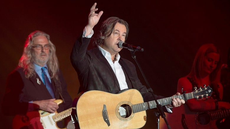 A man holding a guitar and wearing a dark blazer with a white shirt sings into a microphone.