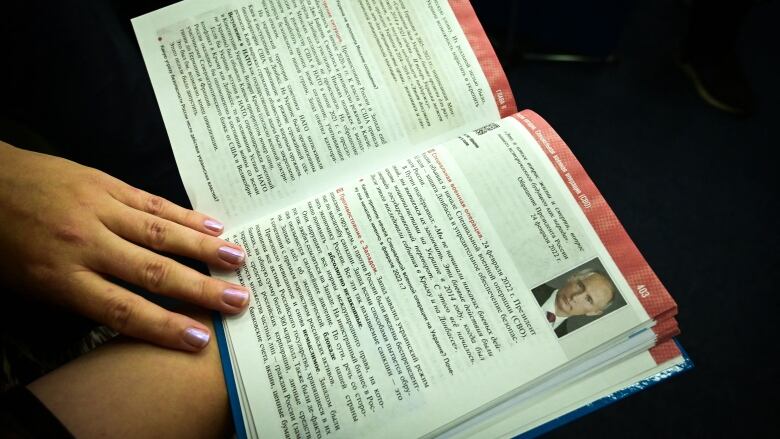 A hand with pink nail polish on the  nails holds open a Russian textbook with a picture of Vladimir Putin inside it.