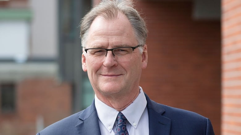 A man in a blue suit with glasses poses for a portrait photograph outside along a railing.