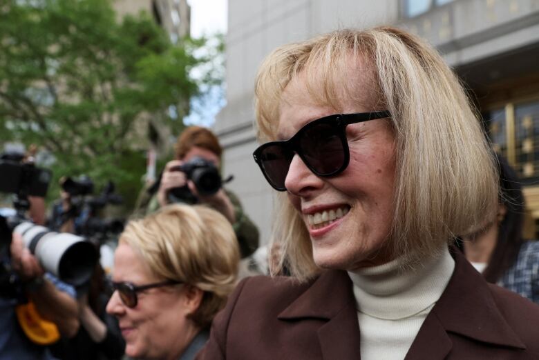 A woman with a blonde bob wears a brown blazer over a white turtleneck and large black sunglasses as she leaves a courthouse surrounded by cameras.