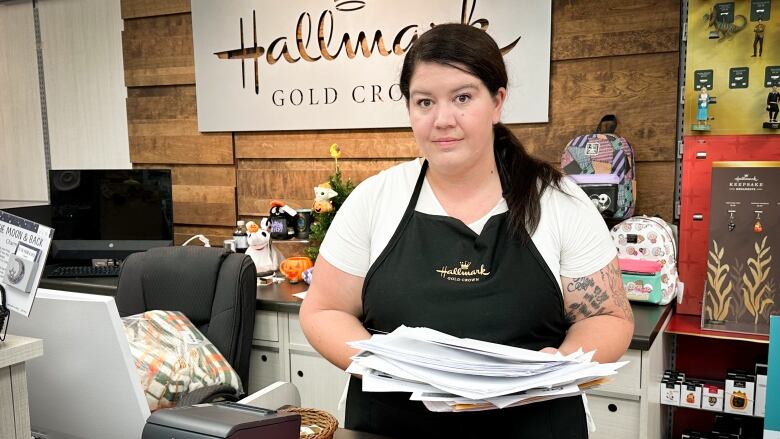 A woman behind a retail counter in a store holds a large sheaf of papers over a red folder labelled resumes.