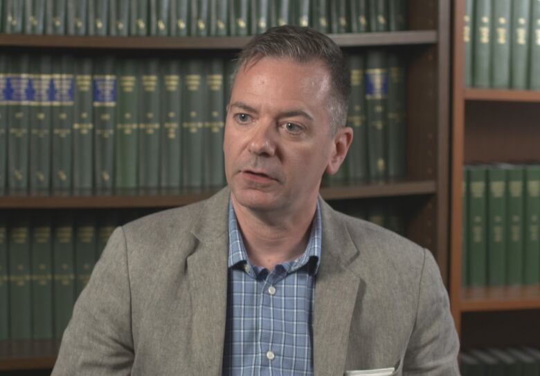 A man in a suit jacket is sitting in front of a bookshelf. 