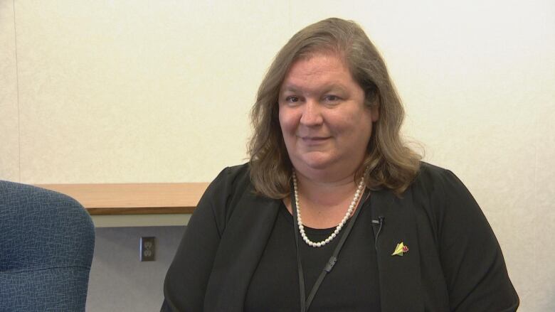 A woman can be seen wearing a black jacket, there is a desk behind her. 