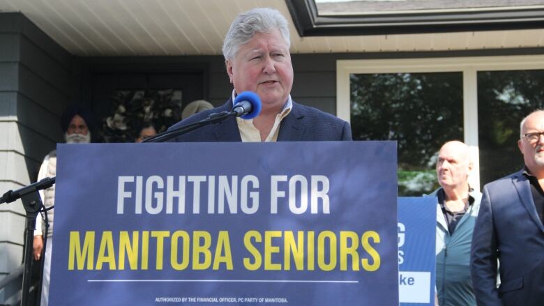 A man in a blue suit speaks at a news conference. The podium has a 'Fighting for Manitoba Seniors' banner on it. 