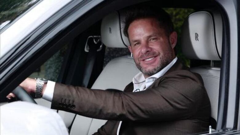 A man smiles at the camera from the driver's seat of a luxurious white car.