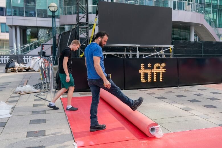 Two men kick out a rolled up red carpet in front of a sign that says TIFF