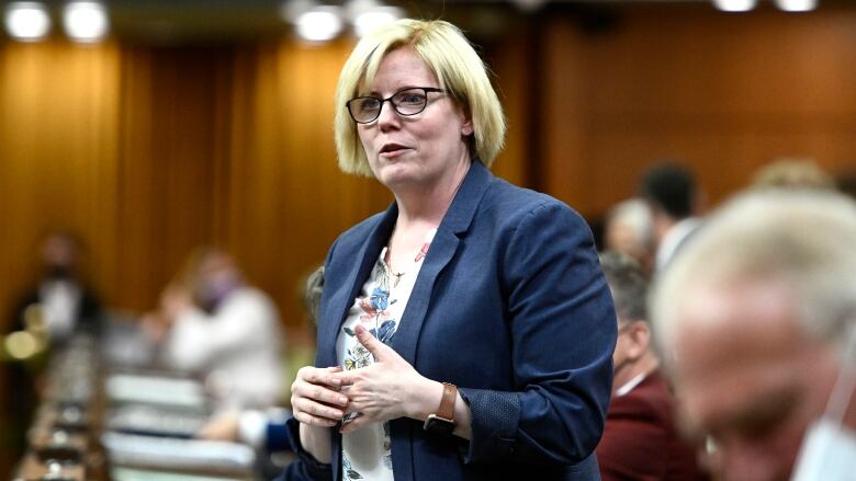 A woman wearing glasses and a blazer stands while speaking in a room filled with people beside and behind her.