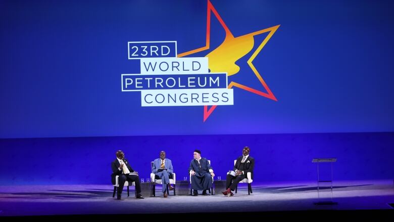 Four men sit in chairs on a stage coloured in purple. On screen behind them is a projection that reads, 23rd World Petroleum Congress.