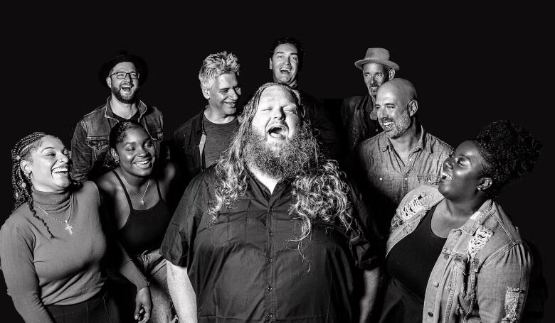 Black and white photo of Matt Anderson and the Big Bottle of Joy: A man with long hair and long beard singing surrounded by three women and five men who all appear to be laughing at him. 