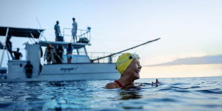 A smiling woman in a swimcap is shown in open ocean by a sailing vessel. 