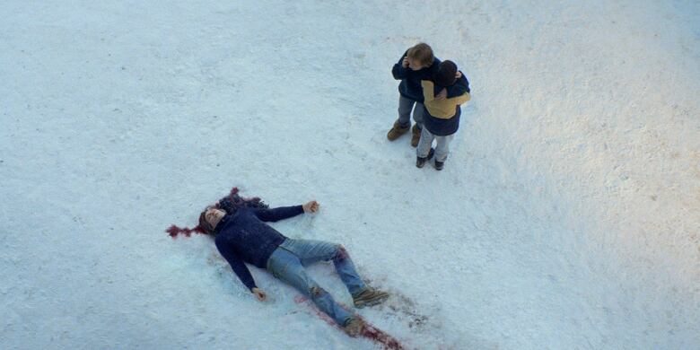 A body lies on its back in the snow, with blood pooling around the head. Nearby, a woman and young boy hold each other. 