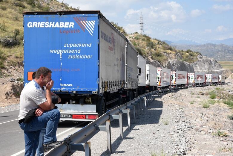 A  man rests behind  a line of transport trucks on a  mountainous road.