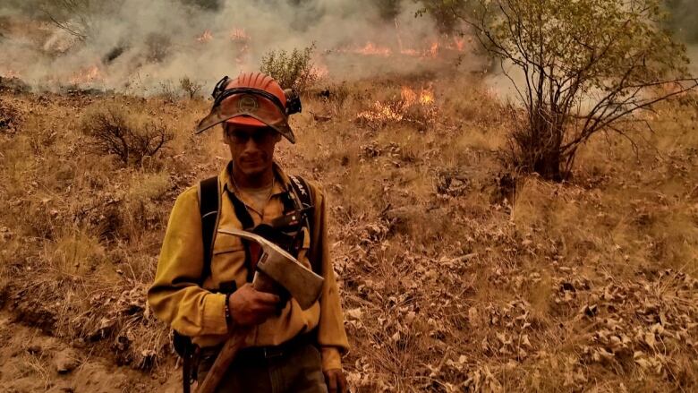 A man with a blackened face in protective gear and holding a fire axe is shown in a dry landscape, with fire and smoke behind him.